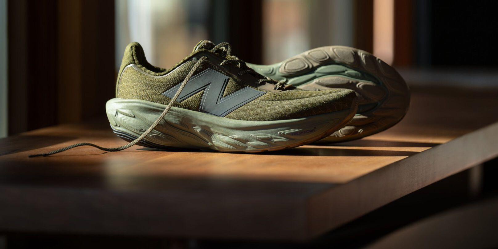Model sitting on wooden and leather chair behind a beige background wearing a tan shirt, yellow hat, and dark New Balance 740s. 